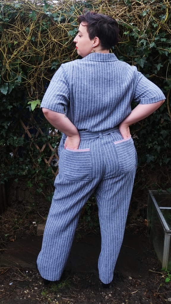 A plus sized woman with short wavy brown hair stands in front of a trellis covered in ivy. She is wearing a grey and white striped jumpsuit. She is turned away from the camera and has her hands in her back pockets.