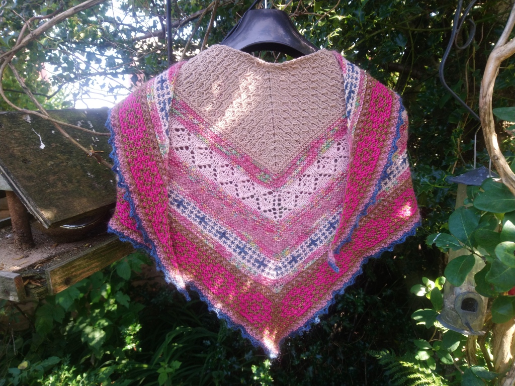 A triangular shawl in seven different yarns is draped on a hanger surrounded my greenery.