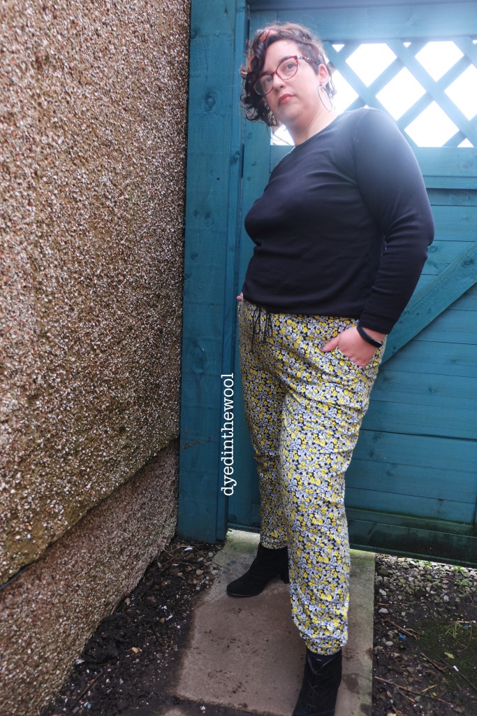 A plus sized woman standing in front of a pebble dashed wall and a blue painted wooden gate, wearing a plain black t-shirt and elasticated cuffed joggers in a very busy floral print. She has her left hand in her pocket and is turned slightly away from the camera.